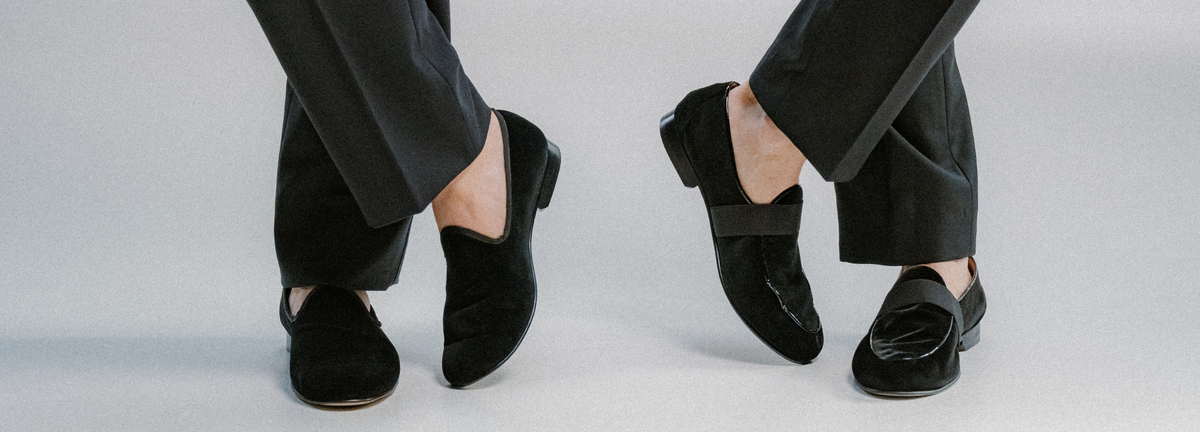A close-up of a person's legs and feet wearing black velvet slip-on loafers with black trousers, photographed against a neutral light gray studio background.