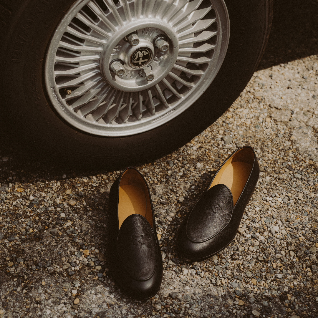 A pair of black leather slip-on loafers with a subtle embossed logo, placed on gravel next to a vintage-style car wheel.